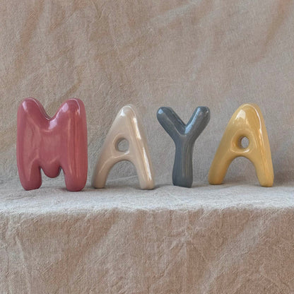 Colorful ceramic letters spelling 'MAYA' on a textured beige surface
